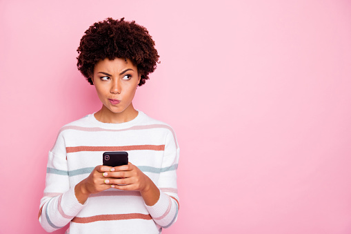 Photo of pretty dark skin lady holding telephone doubtful about new instagram, post wear white striped pullover isolated pastel pink color background