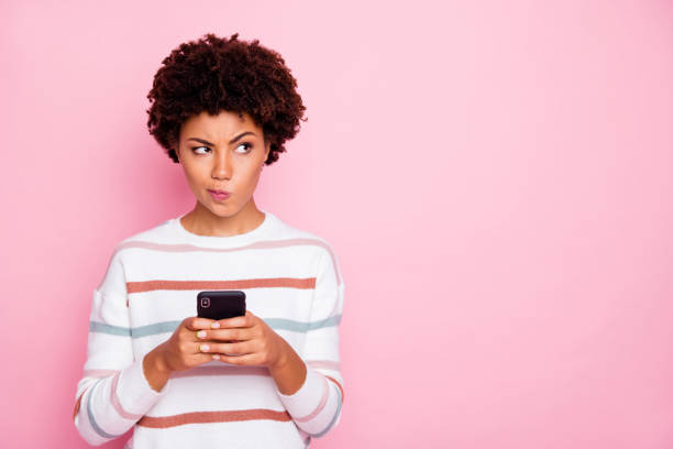 Photo of pretty dark skin lady holding telephone doubtful about new instagram post wear white striped pullover isolated pastel pink color background Photo of pretty dark skin lady holding telephone doubtful about new instagram, post wear white striped pullover isolated pastel pink color background uncertainty photos stock pictures, royalty-free photos & images