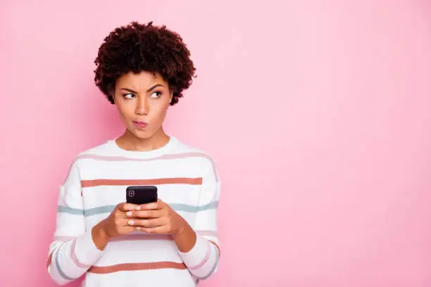 Photo of Photo of pretty dark skin lady holding telephone doubtful about new instagram post wear white striped pullover isolated pastel pink color background
