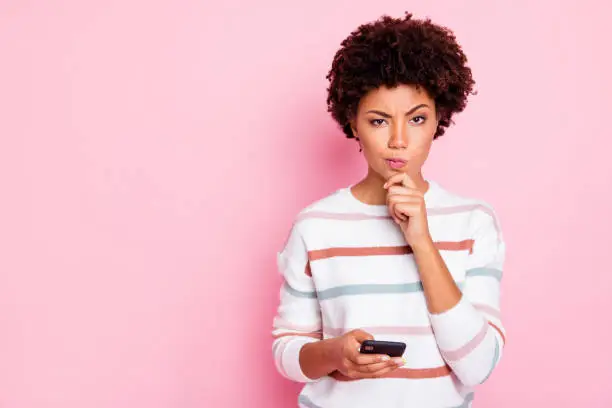 Photo of Photo of pretty dark skin lady holding telephone deciding what comment to write hand touch chin wear white striped pullover isolated pastel pink color background
