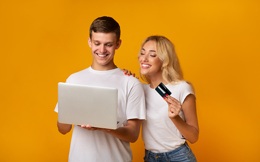 Young couple shopping online with laptop and credit card over yellow studio background, copy space