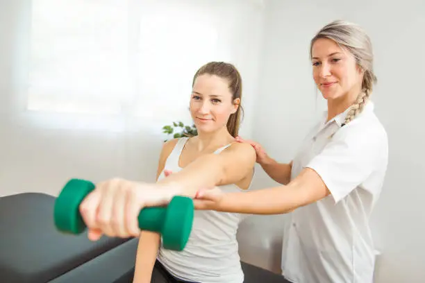 Photo of A Modern rehabilitation physiotherapist at work with client. Working on shoulder with dumbell