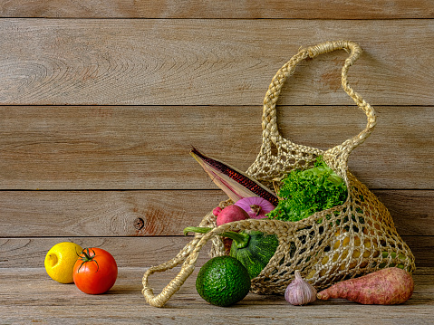 Many colorful contrast color salad vegetables in a reusable string hemp bag on an old weathered wood table against an old weathered wood-paneled rustic background wall, good copy space to the top left of the image.