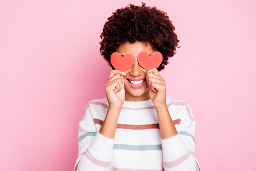 Photo of pretty dark curly skin lady holding in hands little red paper, hearts hiding flirty eyes wear white striped pullover isolated pastel pink color background