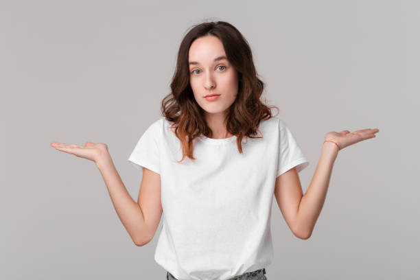 istock Pretty brunette woman in a white tee shrugging her shoulders beacause she is indignant  standing isolated over grey background. Copy space of advertising. 1178555335