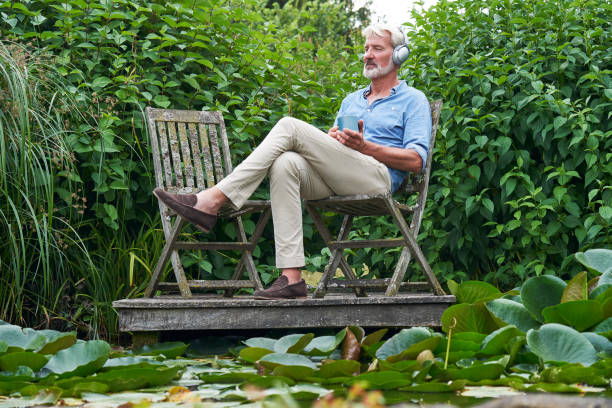 homme mûr détendant dans le jardin écoutant la musique sur des écouteurs sans fil sur la jetée par lac - the thinker audio photos et images de collection