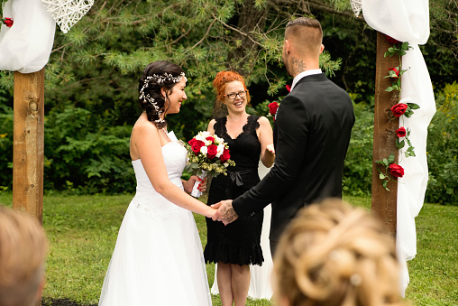 Millennial couple with kids and tattoos saying yes in an intimate outdoors traditional wedding. Here the couple holding hands during the ceremony with woman celebrant. Horizontal full length outdoors shot with some copy space.