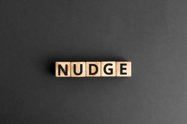 Nudge - word from wooden blocks with letters, pushing gently concept, random letters around, top view gray background
