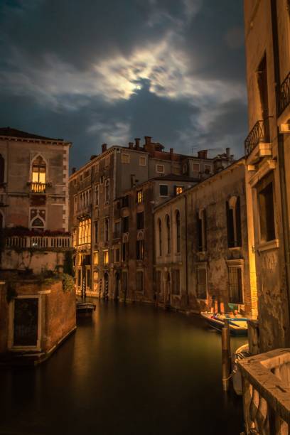 A canal in Venice, Italy during the night with the moon behind clouds and some enlightened windows