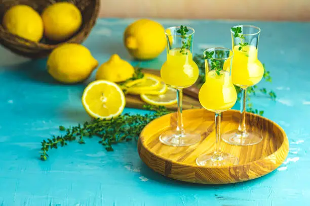 Limoncello with thyme in three grappas wineglass in wooden tray, fresh lemon in basket on light concrete table. Artistic still life on light background.