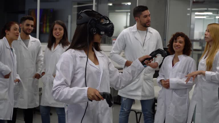 Student wearing a virtual reality headset and holding joysticks experimenting while other students comment and observe during a lab class