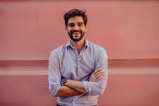 Young man standing by pastel wall and posing with arms crossed looking at camera and smiling