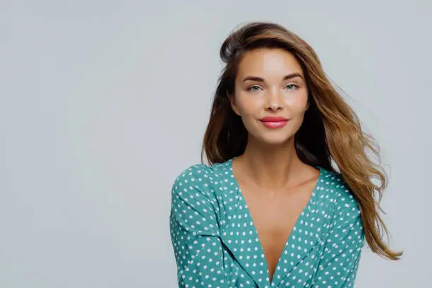 Photo of Studio shot of positive young woman has long wavy hair, makeup, wears turquoise polkadot blouse, looks straightly at camera, models against white background, copy space for your advertisement