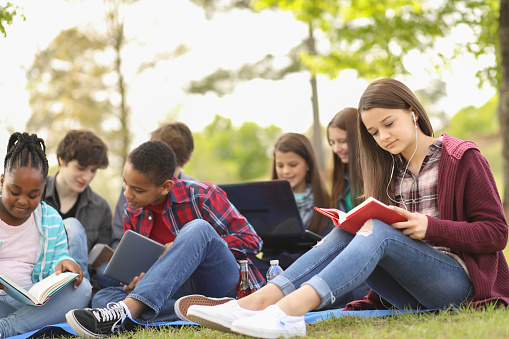 Pre-teenage and teenage group of boys and girls studying, hanging out together in local park or school campus with friends.  Teen girl reading in foreground.