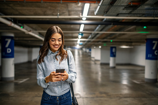 Businesswoman in Underground parking with smartphone. Young woman smiling confident at underground parking lot around cars and lights. Woman in the underground car parking