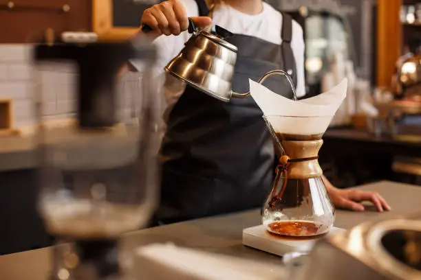 Professional barista preparing coffee using glass coffeemaker pour over coffee maker and drip kettle. Young woman making coffee. Alternative ways of brewing coffee. Coffee shop concept.