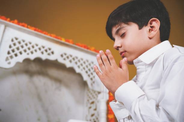 One praying Indian Asian boy doing Pooja or prayer with closed eyes and folded hands in front of a marble temple mandir with a garland of marigold flowers in an intricate carved marble temple mandir