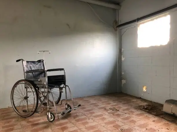 Photo of An empty wheelchair is in an old abandoned room with dirty tiled floor, sunlight shining through ruin window. (Background)