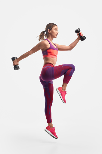 Full body view of muscular young female in sportswear swinging arms with heavy dumbbells and leaping against white background during training for biceps and legs