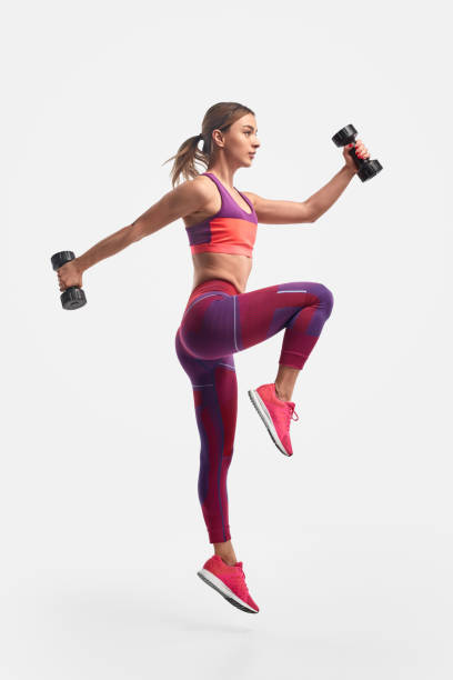 Athletic woman jumping with dumbbells Full body view of muscular young female in sportswear swinging arms with heavy dumbbells and leaping against white background during training for biceps and legs athleticism photos stock pictures, royalty-free photos & images