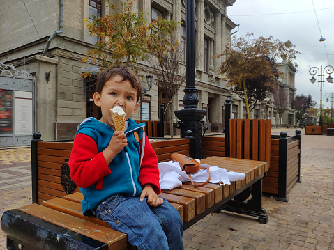 A little boy in a red-blue sweatshirt sits on a bench and eats ice cream on a streetin the city in the summer.