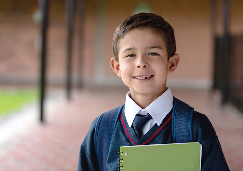 Portrait of a boy at the school holding a notebook in his uniform and looking at the camera smiling - education concepts