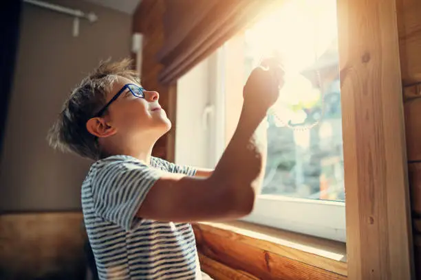 Little boy opening the roller blinds in the morning on sunny day.