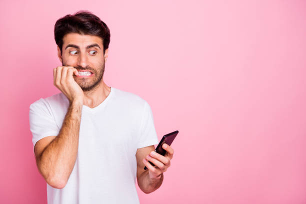 istock Portrait of unhappy scared middle eastern man indian addicted guy use hold cellphone get failure mistake dislike blog notification bite nail wear trendy white t-shirt isolated pastel color background 1178712776