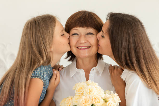 grand-mère retenant des fleurs dans des mains, sa fille et sa petite-fille étreignent l'un l'autre et sourient - mothers day mother kissing child photos et images de collection