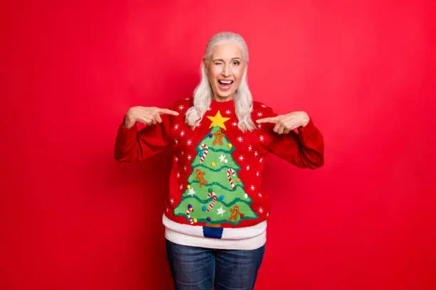 I like my jumper concept. Photo of blinking grey-haired positive hinting, cheerful nice granny showing you her best with small pompons gingerbread men decor pullover isolated bright color background