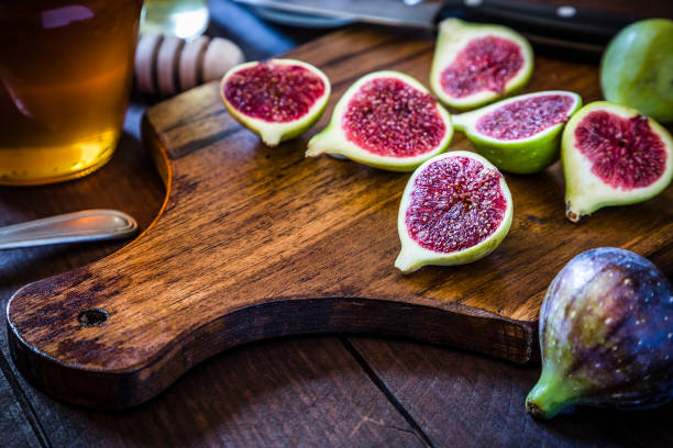 Front view of a rustic cutting board with various sliced figs on top and surrounded by a honey jar, a honey dipper and a kitchen knife. Low key DSLR photo taken with Canon EOS 6D Mark II and Canon EF 24-105 mm f/4L