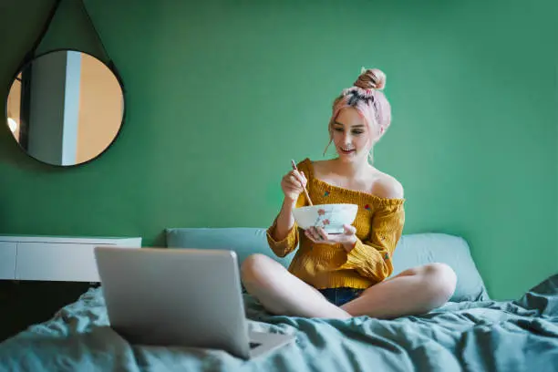 Full length shot of an attractive young woman eating a bowl of pasta and using her laptop in her bedroom