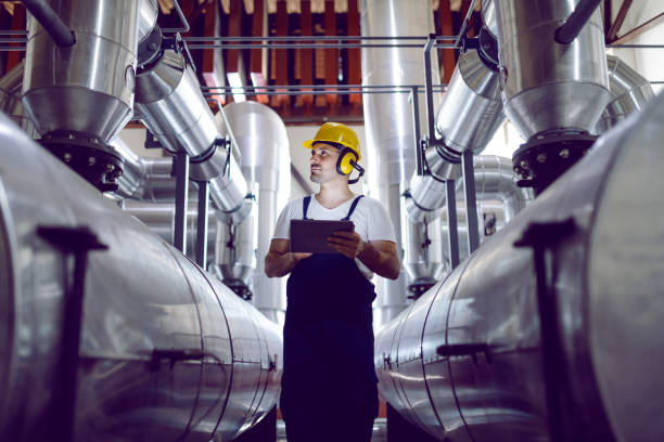 fokussierter pflanzenarbeiter in overalls, mit schutzhelm auf dem kopf und antiphons an den ohren mit tablet te zur kontrolle maschinell. - schwerindustrie stock-fotos und bilder