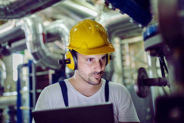 istock Focused plant worker in overalls, with protective helmet on head and antiphons on ears using tablet for checking machine. 1178740099
