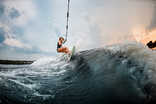 Young sporty girl rides on a green and orange wakeboard in the river near forest