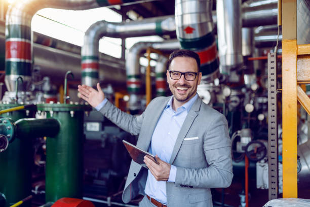 sorridente proprietario caucasico di pianta energetica vestito in abito in possesso di tavoletta e mostrando intorno alla sua pianta. - oil distribution foto e immagini stock