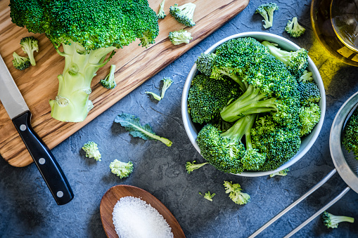 Top view of a bowl full of broccoli branches surrounded by a wooden cutting board with a entire broccoli and a kitchen knife on top, an olive oil bottle, a metal colander and a little wooden tray with salt on a grey textured backdrop. Low key DSLR photo taken with Canon EOS 6D Mark II and Canon EF 24-105 mm f/4L