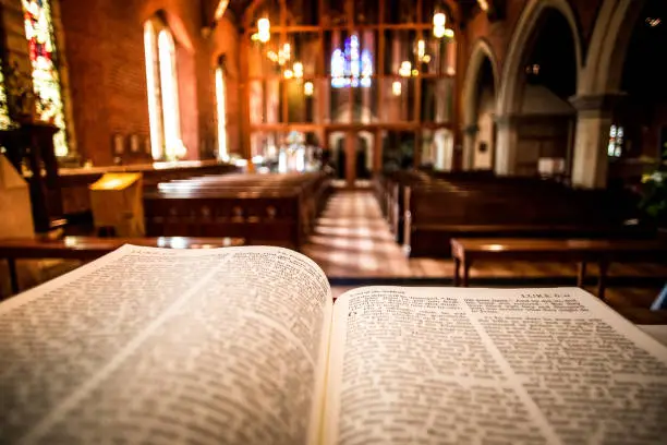 Close up color image depicting the Holy Bible book open on an altar inside an illuminated Anglican church. The book is open to one of the passages of the gospels. Focus is on the book in the foreground, while the interior architecture of the church is defocused beyond, with wooden pews and stained glass windows receding into the distance. Room for copy space.