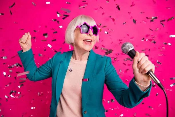 Photo of old mature stylish energetic woman singing in microphone, wearing star shaped spectacles standing in falling confetti isolated over pink vivid color background