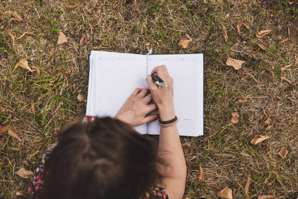 Young girl hides her face with a book lying on a green lawn in the park. Summer day.