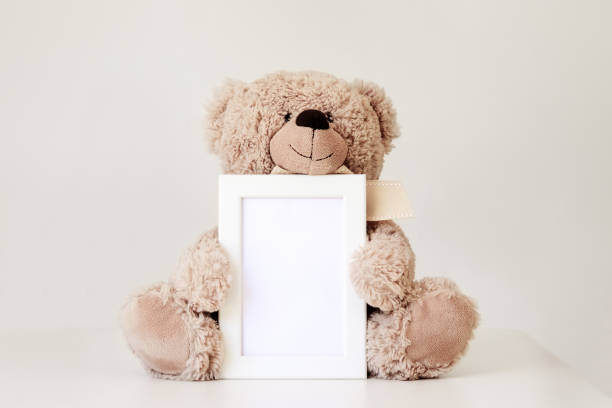 mockup. morbido giocattolo orsacchiotto beige che tiene la cornice mock up bianca pulita con spazio di copia seduto su sfondo grigio chiaro. spazio vuoto. concetto di bambini piccoli. - teddy bear baby toy stuffed animal foto e immagini stock