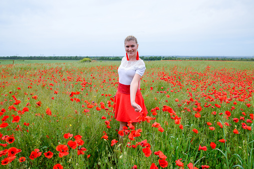Blonde young woman in a red skirt and white shirt, red earrings is in the middle of a poppy field.
