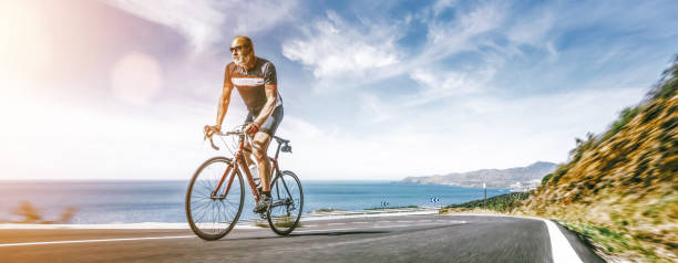 adulte mûr sur un vélo de course escaladant la colline à la route côtière de paysage de mer méditerranéenne - cycling senior adult sports helmet men photos et images de collection
