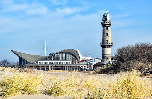 Lighthouse of Warnemünde (Germany) behind sand dunes covered with marram grass, seen from the public beach. Next to the lighthouse there is a remarkable circular building on the beach promenade. It was built in 1968 and houses restaurants and shops.