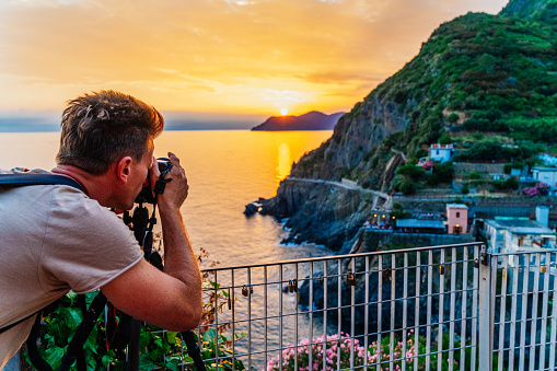 Rear view shot of a photographer taking a picture on seashore under a moody sky at sunset, Cinque Terre, Liguria, Italy