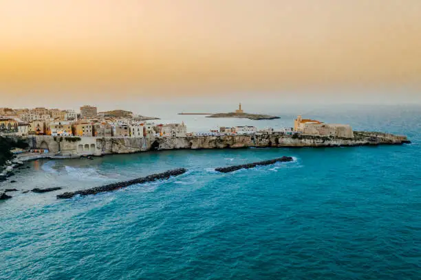 Aerial view of old buildings in a scenic old town on a small peninsula on coastline under a clear orange sky during sunset, Vieste, Puglia, Italy