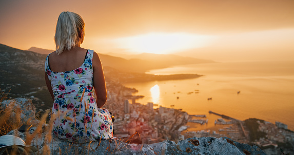 Rear view of a blonde female tourist in a summer dress with floral pattern sitting in mountains and looking at the view of a coastal city during beautiful sunset with orange sky, Monte Carlo, Monaco