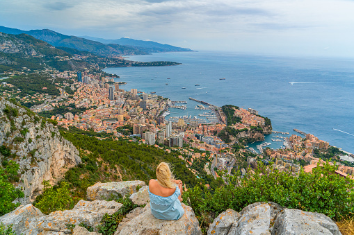 Rear view of a blonde female tourist in a blue summer dress sitting on a rock in mountains and looking at the view of a coastal city during daytime, Monte Carlo, Monaco