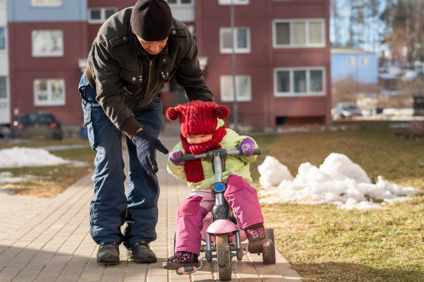 70-years-old homem ativo, avô, ensinando sua neta, menina de 2 anos de idade, como andar de bicicleta durante a caminhada no dia de inverno frio e ensolarado. - playground snow winter little girls - fotografias e filmes do acervo