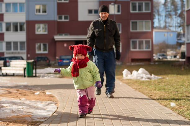 2-летняя девочка бежит на открытом воздухе по дорожке, когда его 70-летний дедушка ходит в фоне. - playground snow winter little girls стоковые фото и изображения